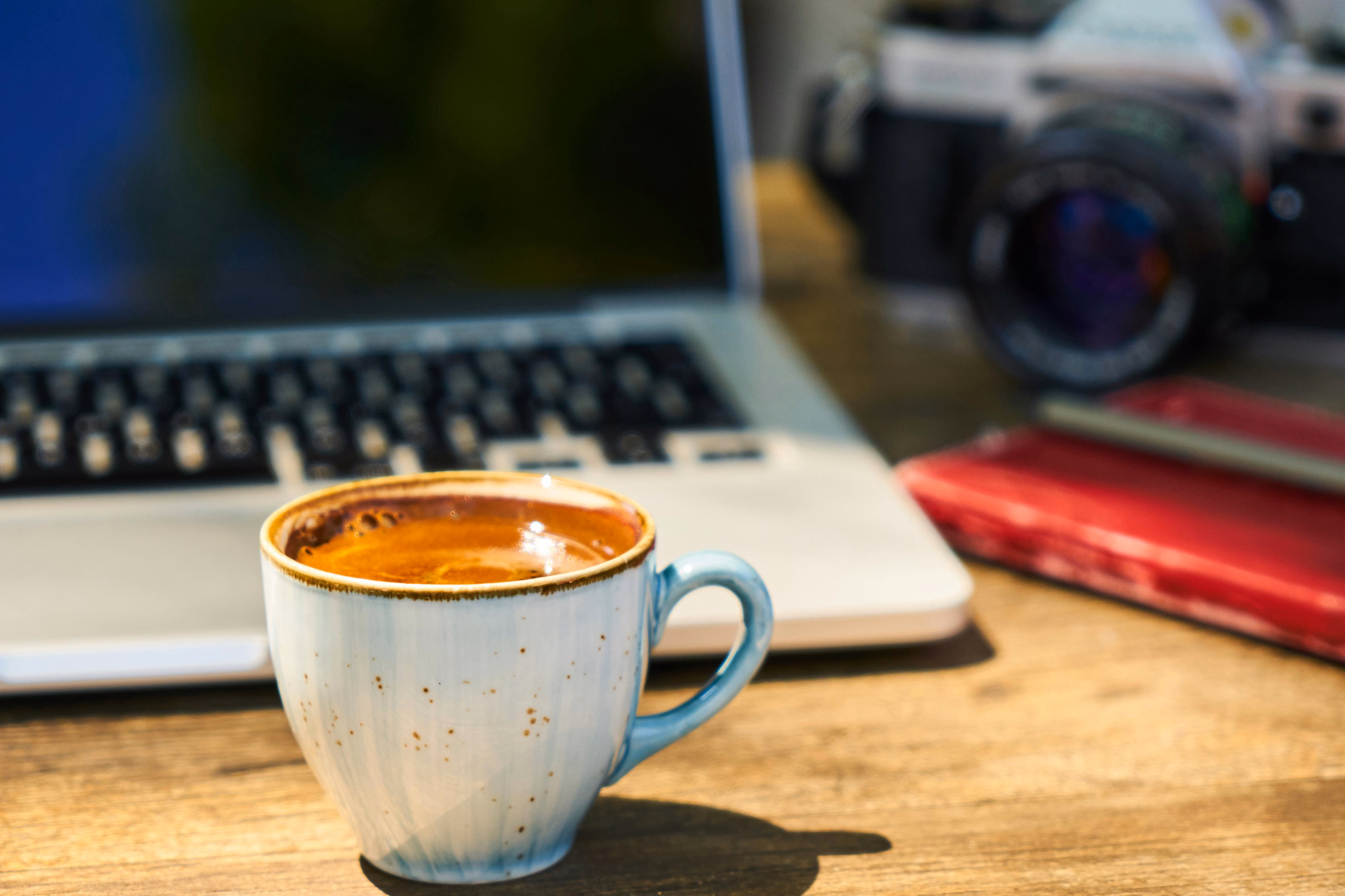 A cup of coffee sits on a wooden desk in front of a laptop, with a camera and red notebook in the background.