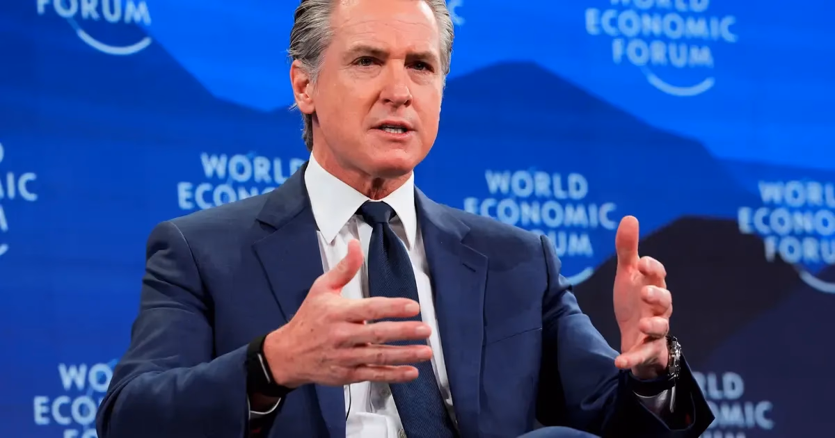 Gavin Newsom, wearing a blue suit and tie, gestures with both hands while speaking in front of a blue backdrop with the words "World Economic Forum" visible.