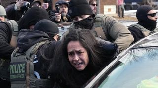 A woman with long dark hair is being arrested by multiple law enforcement officers. She is being pushed into the back of a dark-colored car, and her mouth is open as if she is yelling. The officers are wearing tactical vests with "POLICE FEDERAL OFFICER" printed on the back. Several people are standing around taking pictures.
