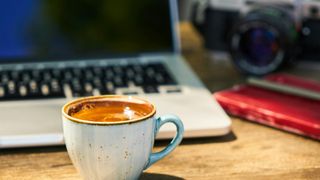 A cup of coffee sits on a wooden desk in front of a laptop, with a camera and red notebook in the background.
