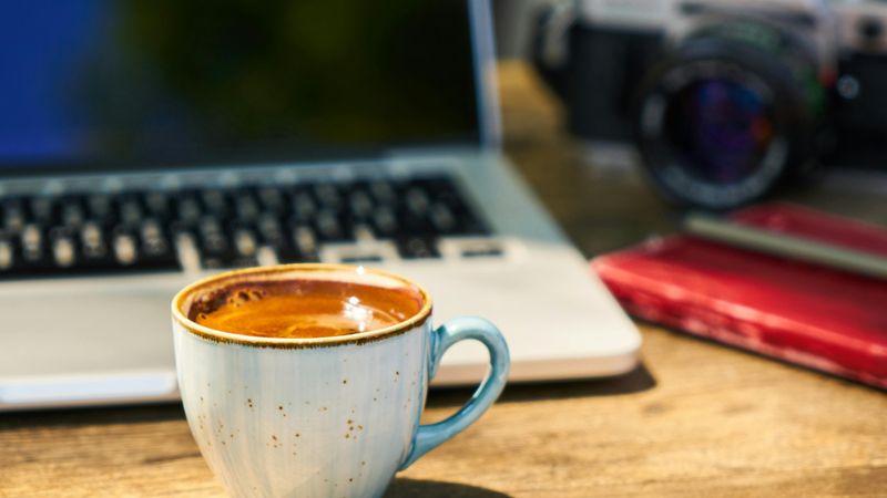 A cup of coffee sits on a wooden desk in front of a laptop, with a camera and red notebook in the background.