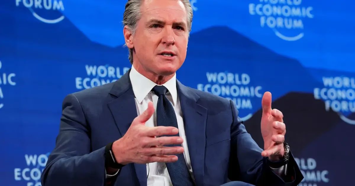 Gavin Newsom, wearing a blue suit and tie, gestures with both hands while speaking in front of a blue backdrop with the words "World Economic Forum" visible.