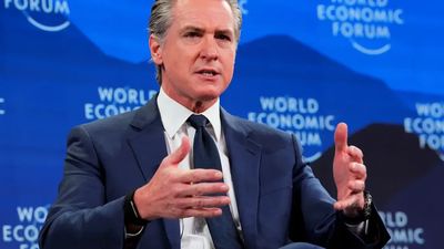 Gavin Newsom, wearing a blue suit and tie, gestures with both hands while speaking in front of a blue backdrop with the words "World Economic Forum" visible.