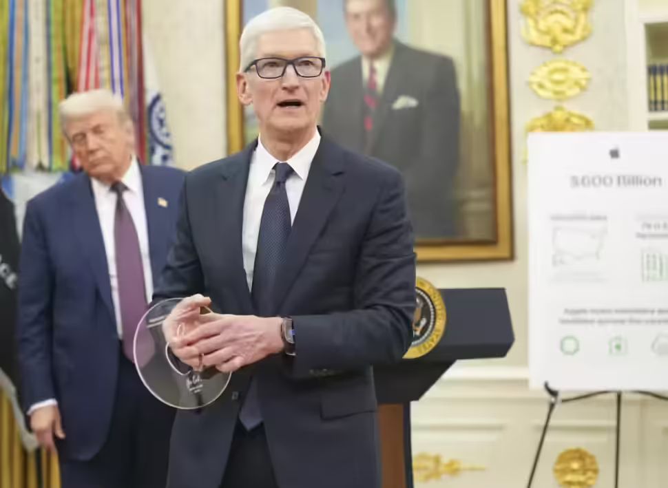 Tim Cook, wearing a dark suit and glasses, holds a clear disc-shaped award while standing at a podium with the Seal of the President of the United States. Donald Trump stands behind him to the left, and a portrait hangs on the wall behind Cook. An easel to the right displays a poster with the text "$600 Billion."