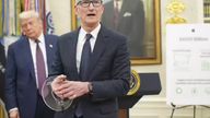 Tim Cook, wearing a dark suit and glasses, holds a clear disc-shaped award while standing at a podium with the Seal of the President of the United States. Donald Trump stands behind him to the left, and a portrait hangs on the wall behind Cook. An easel to the right displays a poster with the text "$600 Billion."