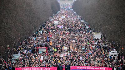 A large crowd of people fills a wide street lined with bare trees, stretching towards the Brandenburg Gate in the distance. Two pink banners in the foreground read, "WIR SIND DIE BRANDMAUER - KEINE ZUSAMMENARBEIT MIT DER AFD!"